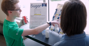 Two individuals stand by a display about sugar content in beverages. One person points at the exhibit while holding a soda can, and the other listens attentively. Various props and posters are visible.